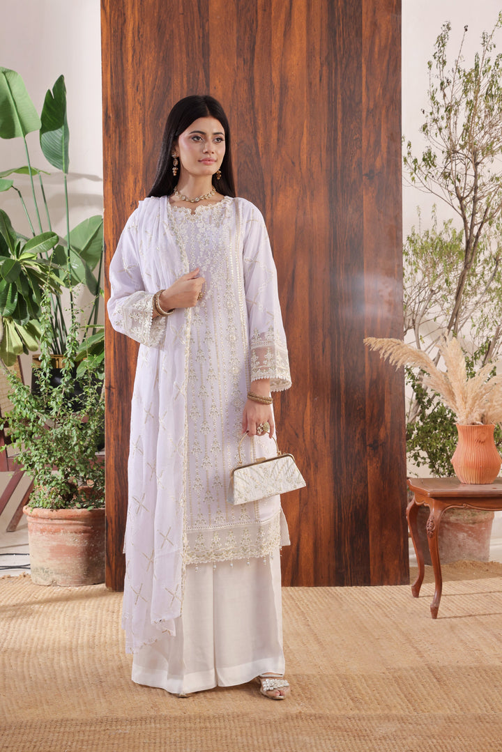 RIWAYAT - Woman in a white traditional outfit standing in a room with plants and wooden elements.