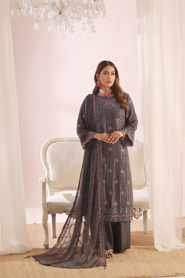 Woman in a dark gray traditional outfit with intricate patterns standing in a decorated room.