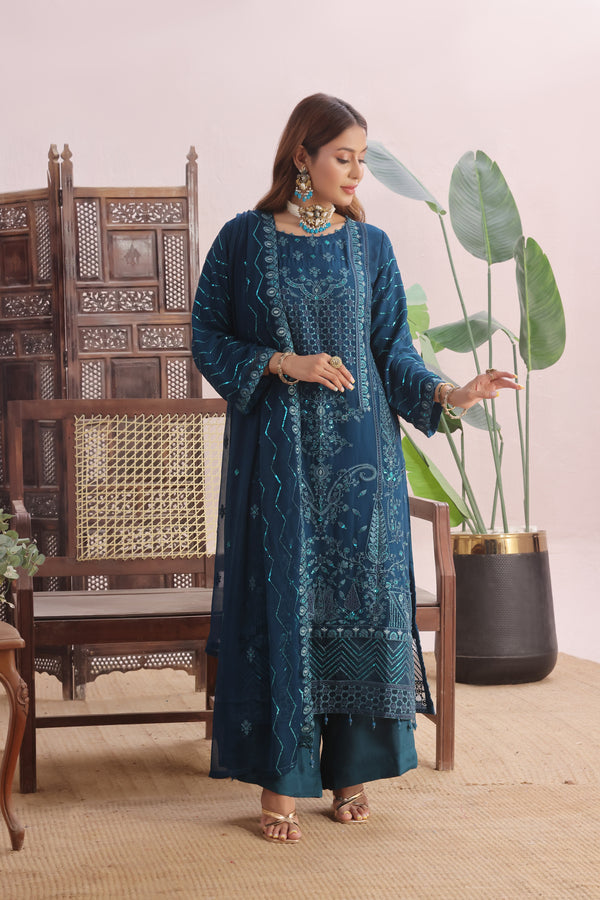 Woman in a blue embroidered outfit standing in a room with a plant and wooden chair.